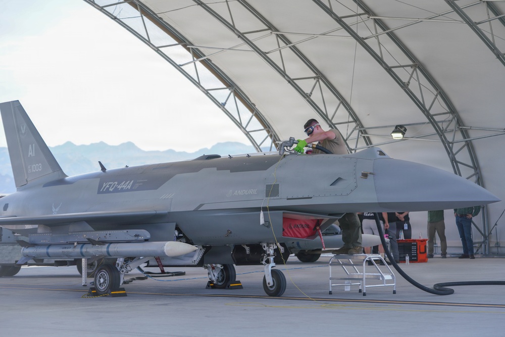 A gray military aircraft designated 'YFQ-44A' with Anduril branding and tail number 1003 is parked under an open-sided hangar structure. A technician is working in the cockpit area while ground crew members stand nearby. The aircraft appears to be undergoing pre-flight or maintenance servicing, with a fuel hose connected and munitions visible under the wings.