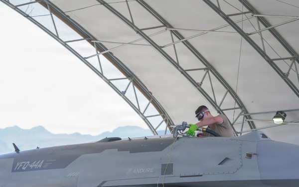 A gray military aircraft designated 'YFQ-44A' with Anduril branding and tail number 1003 is parked under an open-sided hangar structure. A technician is working in the cockpit area while ground crew members stand nearby. The aircraft appears to be undergoing pre-flight or maintenance servicing, with a fuel hose connected and munitions visible under the wings.