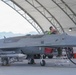A gray military aircraft designated 'YFQ-44A' with Anduril branding and tail number 1003 is parked under an open-sided hangar structure. A technician is working in the cockpit area while ground crew members stand nearby. The aircraft appears to be undergoing pre-flight or maintenance servicing, with a fuel hose connected and munitions visible under the wings.
