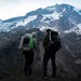 Two backpackers stand on a rocky ridge overlooking a dramatic glaciated mountain landscape, viewed from behind. They are equipped with full hiking packs and appear to be pausing to take in the view of snow-covered peaks and glaciers. The lighting suggests either early morning or late evening conditions.