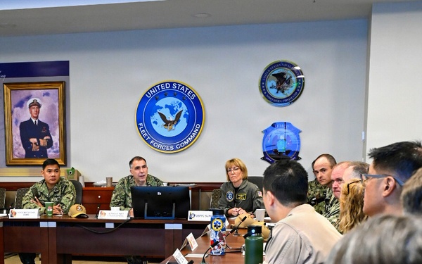 A formal military briefing or meeting is taking place at United States Fleet Forces Command headquarters. Multiple military personnel from various branches are seated around a large conference table, with name placards visible. A female officer in a flight suit appears to be leading or presenting at the head of the table.