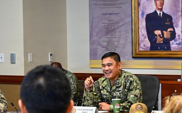 A U.S. Navy meeting or briefing is taking place in a formal conference room. A smiling service member identified by a nameplate as 'Force Master Chief BUMED' is seated at a table, appearing to address attendees. A painted portrait of a senior naval officer hangs on the wall behind the table.
