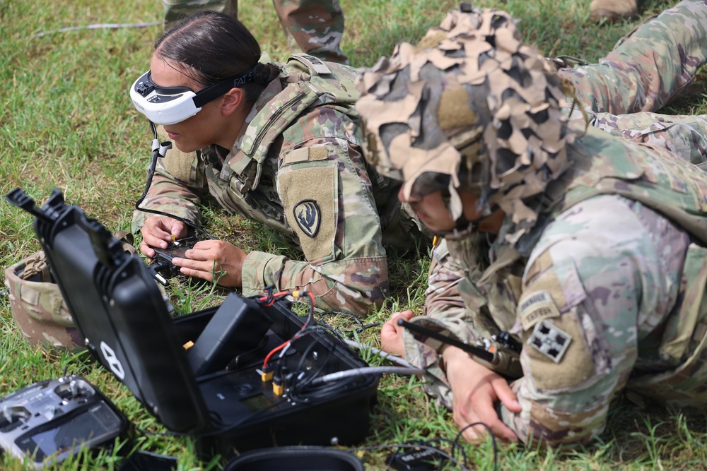 Military personnel in camouflage uniforms are lying prone on grass while operating drone equipment. One soldier is wearing FPV (First Person View) goggles, suggesting active drone piloting, while others assist with what appears to be a small drone or UAS (Unmanned Aerial System) setup. The scene depicts a field training exercise involving drone operations.