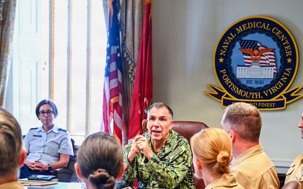 A military briefing or meeting is taking place at Naval Medical Center Portsmouth, Virginia (established 1830), as indicated by the official seal on the wall. A senior officer in camouflage utilities is addressing a group of personnel seated around a conference table, most wearing tan/khaki service uniforms. One individual in the background appears to be wearing an Air Force service uniform, suggesting an inter-service gathering.