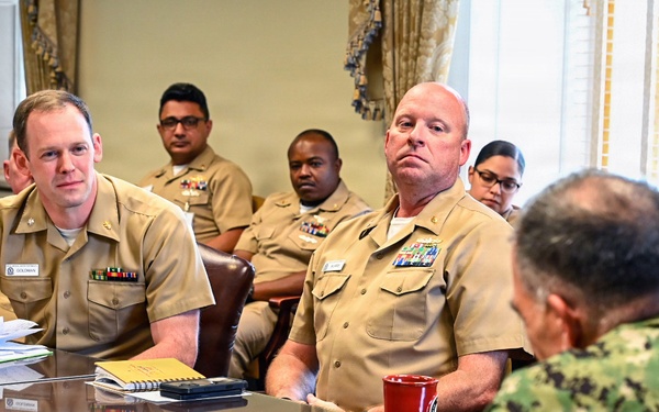 A formal military briefing or meeting is taking place around a large reflective conference table. Multiple U.S. Navy personnel in khaki service uniforms are seated and listening to a speaker in Navy Working Uniform (NWU camouflage pattern) whose back is to the camera. The setting appears to be an official government or military headquarters conference room with formal decor.