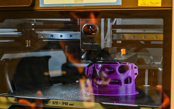A Bambu Lab 3D printer is actively printing a purple lattice/mesh-structured object on its build plate. The printer's enclosed chamber is visible through a glass door, with the print head positioned above the partially completed purple filament structure. The build volume dimensions '340 × 320 × 340 mm³' are visible on the front panel.