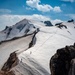 A high-altitude mountaineering scene showing a snow-covered glacial slope flanked by dark volcanic or rocky terrain. At least two small human figures are visible traversing the snowfield, illustrating the vast scale of the mountain. A prominent snow-capped peak rises in the background under a partly cloudy blue sky.