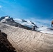 Two mountaineers are traversing a snow and ice field on a high-altitude mountain under a clear blue sky. The foreground climber carries a large pack with an orange cover and uses trekking poles, while a second climber is visible further along the route. A prominent rocky summit with glaciated slopes dominates the background.