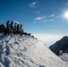 A group of approximately five to six mountaineers stand silhouetted on a snow-covered summit ridge under a bright sunstar in a deep blue sky. The climbers are equipped with helmets, backpacks, and mountaineering gear, suggesting a technical alpine ascent. A jagged rocky peak is visible in the background, partially shrouded in clouds below the summit level.