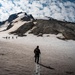 A group of mountaineers ascends a snow-covered volcanic peak under a partly cloudy sky. The foreground features a lone climber with a backpack and trekking pole, while several others are visible further up the slope. The mountain displays a mix of snow fields and dark volcanic rock formations.