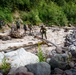 A group of hikers with backpacks and trekking poles are navigating a rocky, fast-moving glacial river crossing using fallen logs as makeshift bridges. The terrain is rugged, with large boulders, driftwood, and turbulent milky-white water characteristic of glacial melt. Dense green forest covers the hillside in the background.