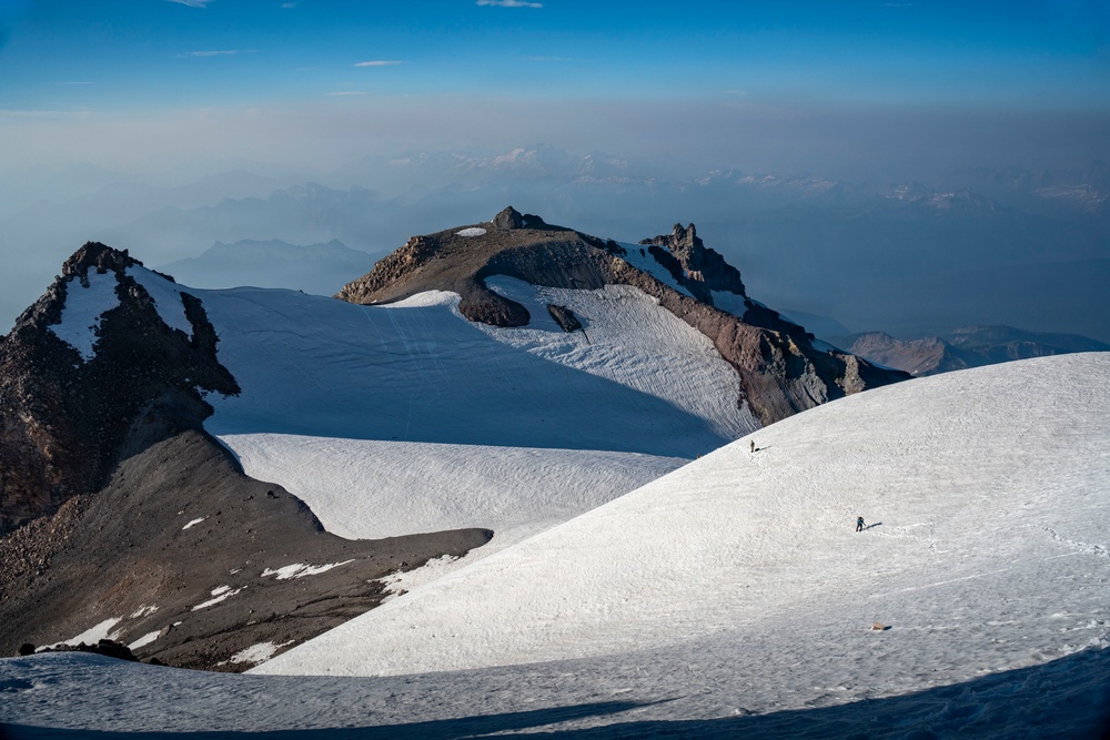 A high-altitude mountain landscape featuring expansive snow and glacier fields with rocky volcanic peaks in the background. Two small human figures are visible on the snow slope in the foreground, emphasizing the vast scale of the terrain. The scene is photographed from an elevated vantage point, with a hazy valley and distant mountain ranges visible far below.