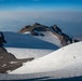 A high-altitude mountain landscape featuring expansive snow and glacier fields with rocky volcanic peaks in the background. Two small human figures are visible on the snow slope in the foreground, emphasizing the vast scale of the terrain. The scene is photographed from an elevated vantage point, with a hazy valley and distant mountain ranges visible far below.