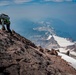 A mountaineer is scrambling up a steep rocky ridge using hands and feet, carrying a large backpack with a helmet attached. In the background, two additional climbers are visible on a snow-covered slope below. The scene features dramatic alpine terrain with glaciated peaks and snow fields extending into the distance under a hazy blue sky.