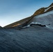 A high-altitude glacial landscape photographed at either dawn or dusk, showing a large snow and ice field on a steep mountain slope. The foreground features heavily striated glacier ice with visible flow lines, while the upper portion reveals rocky outcrops illuminated by warm golden light. Several small human figures are barely visible on a ridge in the middle distance, providing scale.