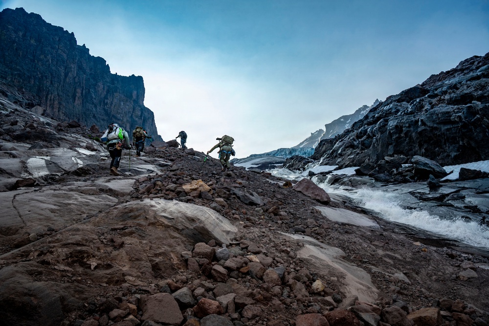 A group of four mountaineers with heavy backpacks ascend a rocky glacial terrain flanked by steep dark cliffs. A rushing glacial meltwater stream runs alongside their path on the right side. The scene depicts a challenging high-altitude expedition under an overcast sky with remnants of snow and ice visible throughout.