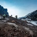 A group of four mountaineers with heavy backpacks ascend a rocky glacial terrain flanked by steep dark cliffs. A rushing glacial meltwater stream runs alongside their path on the right side. The scene depicts a challenging high-altitude expedition under an overcast sky with remnants of snow and ice visible throughout.