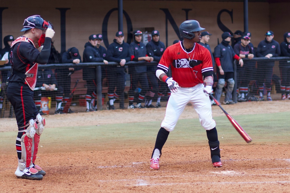 A college baseball game is in progress at home plate, with a batter in a red jersey having just completed a swing while the catcher stands nearby with his hand raised to his helmet. Both players appear to be on the same team based on matching red and black uniforms. A dugout full of teammates in black uniforms watches from behind a protective net in the background.