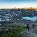 Three hikers with large backpacks are ascending a rocky alpine trail at either dawn or dusk, with warm golden light illuminating distant mountain peaks. Below them lies a striking turquoise glacial lake nestled in a volcanic or glacially-carved basin surrounded by patchy snow fields. The scene depicts a high-altitude wilderness environment with expansive mountain ranges visible in the background.