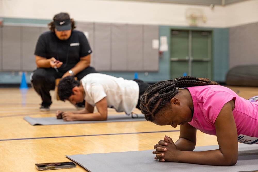 Two children are performing plank exercises on gray yoga mats in what appears to be a school gymnasium. An adult instructor, wearing a black Nike visor and black athletic clothing, crouches in the background observing and timing the exercise using a handheld device. The setting suggests a physical education class or youth fitness program.