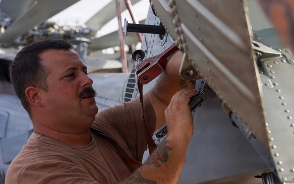 A man with a mustache and tattoos on his forearm is performing maintenance work on what appears to be a military helicopter, likely on the deck of a naval vessel. He is using a tool to work on a mechanical component near the tail rotor or blade assembly of the aircraft. Another person and additional aircraft are visible in the background.