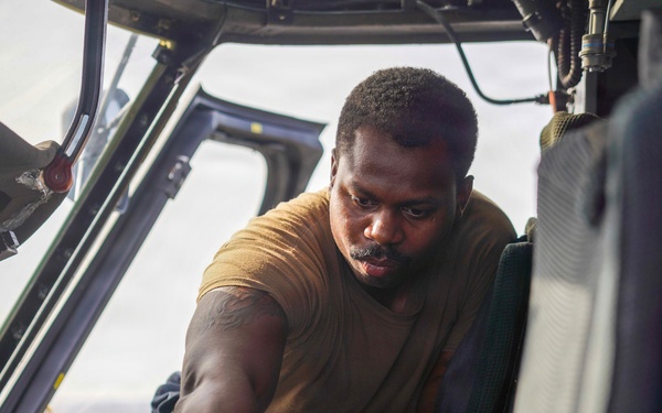 A technician in a tan military-style t-shirt is performing maintenance work inside an aircraft cockpit. He is leaning over the center console, working with gloved hands on avionics or flight control components. The setting appears to be a military or large transport aircraft based on the cockpit configuration and seat design.