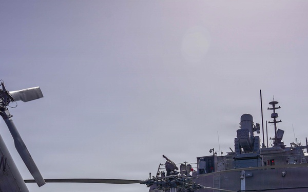 A U.S. Navy helicopter with folded rotor blades is parked on the flight deck of a naval vessel, with crew members in red and dark uniforms observing a second ship alongside. The helicopter bears 'NAVY' markings and tail code 'NW' with partial designation 'HSM-60' visible. Two naval vessels appear to be conducting an underway replenishment or close-proximity maritime operation at sea.