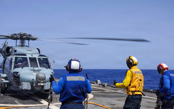 Three naval deck crew members are managing a military helicopter on a ship's flight deck at sea. The crew, wearing color-coded safety gear, are handling tie-down chains while the helicopter's rotors are spinning. The scene depicts a standard naval aviation deck operation, likely a pre-launch or post-landing procedure.