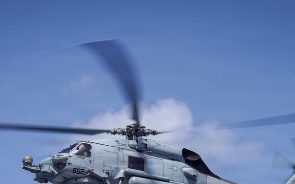 A U.S. Navy MH-60R Seahawk helicopter is captured in flight against a partly cloudy blue sky. The aircraft bears the markings '602' on its nose and 'HSM-60' squadron designation along with 'NAVY' on the fuselage. The helicopter appears to be in a hover or low-speed flight configuration with its main rotor blades visibly blurred from rotation.