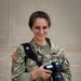 A U.S. Army soldier in OCP (Operational Camouflage Pattern) uniform is smiling and holding a Canon DSLR camera with a 24-70mm lens. She is wearing a camera strap across her body and has a U.S. flag patch on her left shoulder. The setting appears to be outdoors against a stone or concrete wall.