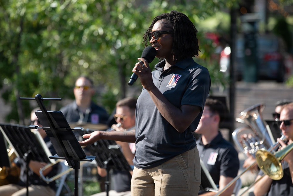 A person wearing a dark navy polo shirt with an embroidered logo is speaking or singing into a handheld microphone at an outdoor event. They are standing in front of a music stand with other band members visible in the background, all wearing matching navy uniforms. The setting appears to be an outdoor public performance, likely a military or civic band concert.