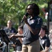 A person wearing a dark navy polo shirt with an embroidered logo is speaking or singing into a handheld microphone at an outdoor event. They are standing in front of a music stand with other band members visible in the background, all wearing matching navy uniforms. The setting appears to be an outdoor public performance, likely a military or civic band concert.