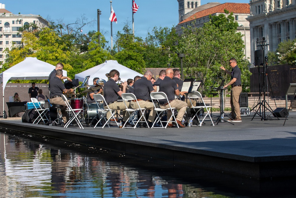 A uniformed ensemble, likely a military or law enforcement band, performs an outdoor concert on a low stage adjacent to a reflective pool. The musicians, dressed in matching dark polo shirts and khaki pants, are directed by a conductor standing at a podium on the right. The setting appears to be a public memorial or civic plaza in an urban environment, with flags, white event tents, and a classical-style building visible in the background.