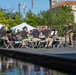A uniformed ensemble, likely a military or law enforcement band, performs an outdoor concert on a low stage adjacent to a reflective pool. The musicians, dressed in matching dark polo shirts and khaki pants, are directed by a conductor standing at a podium on the right. The setting appears to be a public memorial or civic plaza in an urban environment, with flags, white event tents, and a classical-style building visible in the background.