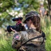 A female National Guard soldier is photographed from behind while operating a Canon DSLR camera equipped with a Rode microphone, suggesting a public affairs or combat camera role. She is wearing OCP (Operational Camouflage Pattern) uniform with a tactical vest clearly labeled 'NATIONAL GUARD' on the back. An American flag patch is visible on her left sleeve, and a partial name tape reading 'PICKI...' is visible on her cap.