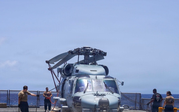 A military helicopter, appearing to be an MH-60 Seahawk, is positioned on the flight deck of a naval vessel at sea. Several crew members in casual and working uniforms are gathered around the aircraft, likely conducting maintenance, inspection, or securing operations. The scene takes place under bright daylight conditions with open ocean visible in the background.