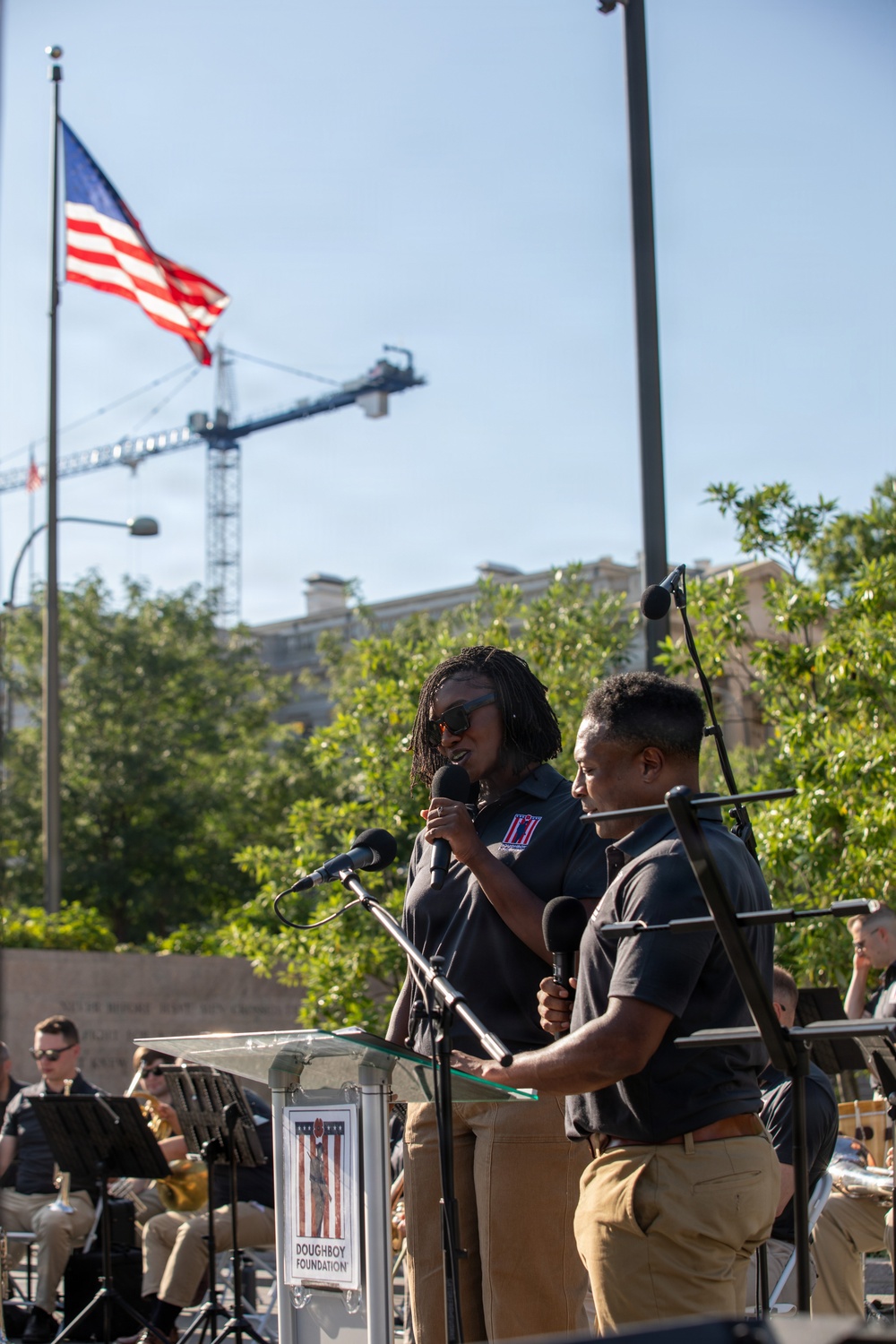 Two individuals in matching black polo shirts and khaki pants are speaking at a podium bearing the 'Doughboy Foundation' logo at an outdoor event. An American flag flies prominently in the background alongside a construction crane and trees. Behind them, a band or ensemble of musicians in similar attire can be seen seated with instruments and music stands.
