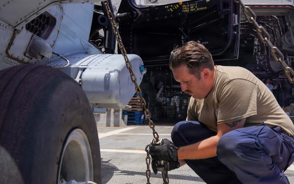 A military or naval aviation technician is crouching on a ship's flight deck, securing a helicopter's landing gear with chains. The worker is attaching or inspecting tie-down chains connected to the aircraft's wheel assembly, which is also blocked with a yellow wheel chock. The setting appears to be an outdoor flight deck of a naval vessel in bright daylight conditions.