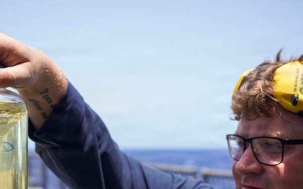 A uniformed military or naval personnel member holds up a glass jar containing a yellowish-green liquid sample while standing on what appears to be the deck of a naval vessel. The individual is wearing glasses, a yellow hearing protection device pushed up on their forehead, and a dark blue uniform with a partially visible name tag. The ocean and blue sky are visible in the background.
