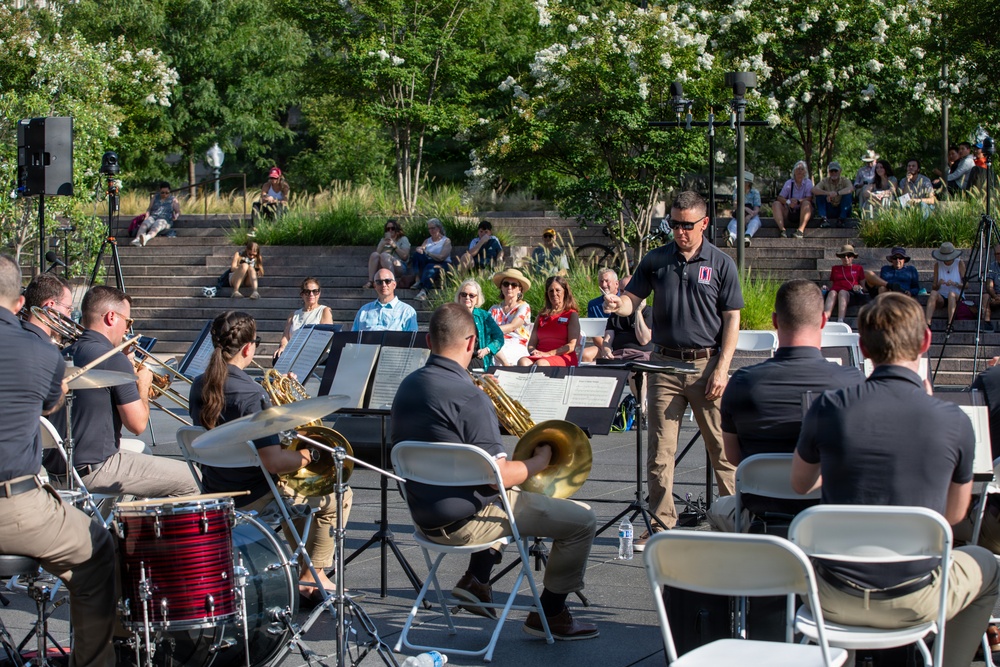 An outdoor band performance is taking place in what appears to be a public park or plaza with amphitheater-style stone seating steps. A conductor in a dark polo shirt with a patch/insignia stands at a music stand directing a brass and percussion ensemble, all dressed in matching dark shirts and khaki pants. Audience members are seated both in folding chairs near the performers and on the stone steps in the background.