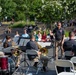 An outdoor band performance is taking place in what appears to be a public park or plaza with amphitheater-style stone seating steps. A conductor in a dark polo shirt with a patch/insignia stands at a music stand directing a brass and percussion ensemble, all dressed in matching dark shirts and khaki pants. Audience members are seated both in folding chairs near the performers and on the stone steps in the background.