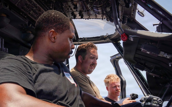 Three individuals are seated inside the cockpit of a large military aircraft, examining the flight controls and instrument panels. The cockpit features complex avionics, control yokes, and overhead panels typical of a large military transport or bomber aircraft. The setting appears to be a static display or ground inspection, as the canopy is open and natural daylight is visible.