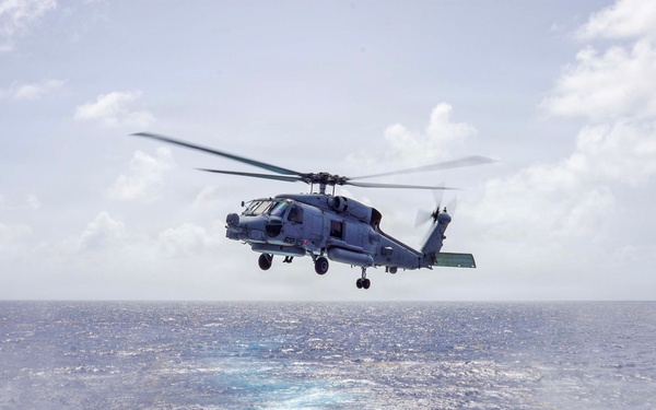 A military helicopter, consistent with an MH-60 Seahawk, is hovering or departing from the flight deck of a naval vessel over open ocean. The rotor wash is visibly churning the water below the aircraft, creating a circular white foam pattern on the sea surface. The scene depicts a maritime helicopter operation, likely a launch or approach to a ship's flight deck.