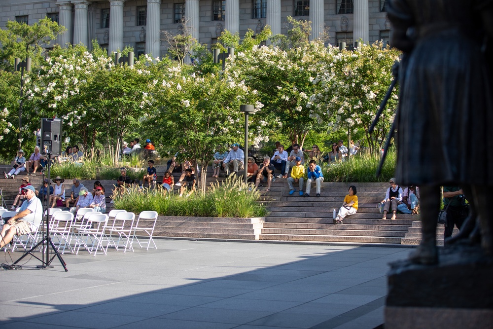 An outdoor public gathering or event is taking place in what appears to be a plaza or public square in front of a large neoclassical government or civic building. Attendees are seated on tiered stone steps and white folding chairs arranged in rows, suggesting a performance or ceremony is underway. A bronze statue is partially visible in the foreground right, and blooming white-flowered trees provide a lush backdrop.