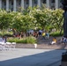 An outdoor public gathering or event is taking place in what appears to be a plaza or public square in front of a large neoclassical government or civic building. Attendees are seated on tiered stone steps and white folding chairs arranged in rows, suggesting a performance or ceremony is underway. A bronze statue is partially visible in the foreground right, and blooming white-flowered trees provide a lush backdrop.