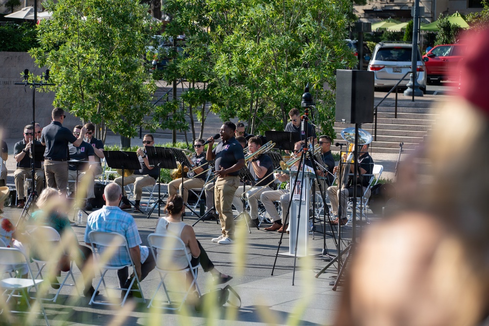 An outdoor band performance is taking place in what appears to be a public plaza or courtyard during daytime. A vocalist holding a microphone stands at the center while a large ensemble of musicians seated in chairs play brass and wind instruments behind him. A small audience watches from folding chairs in the foreground.