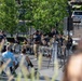 An outdoor band performance is taking place in what appears to be a public plaza or courtyard during daytime. A vocalist holding a microphone stands at the center while a large ensemble of musicians seated in chairs play brass and wind instruments behind him. A small audience watches from folding chairs in the foreground.