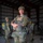 A military paratrooper in full jump gear stands confidently with arms crossed inside a hangar, wearing OCP (Operational Camouflage Pattern) uniform and a tan ballistic helmet. The subject has a visible prosthetic leg below the right knee, decorated with colorful stickers including 'EVO' and 'JUST CHOP IT' text. Other soldiers in similar parachute equipment are visible in the background preparing for a jump operation.