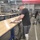 A man with a prosthetic lower leg is working at a long wooden workbench in what appears to be a military parachute rigging facility. He is handling parachute lines and canopy material, with numerous packed parachute bags visible on shelves and in wire carts throughout the facility. An American flag is partially visible in the upper right corner, and two other workers are visible in the background.