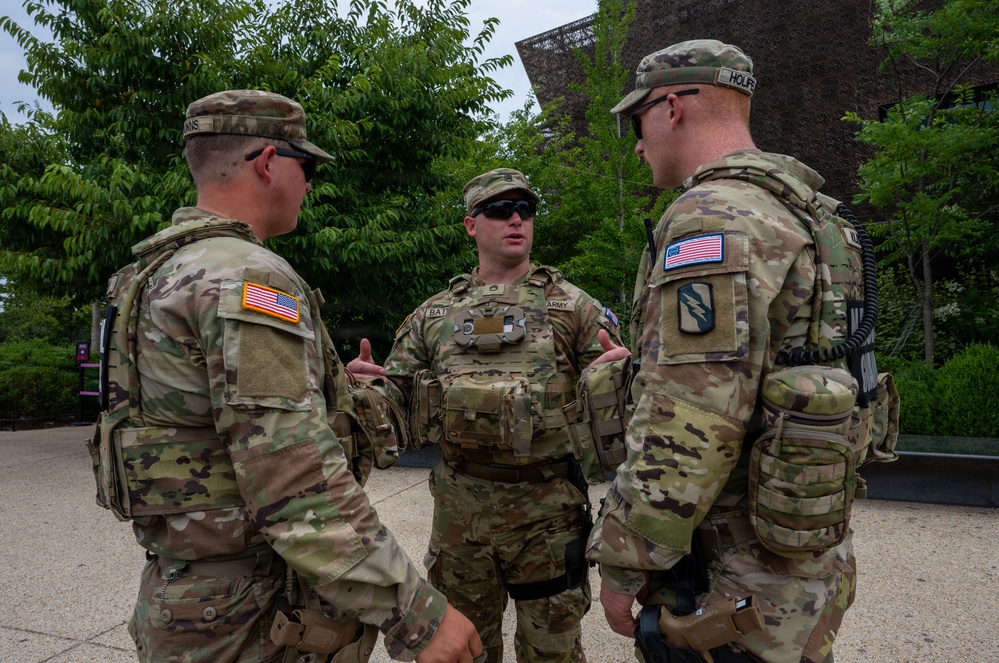 Three U.S. Army soldiers in Operational Camouflage Pattern (OCP) uniforms are engaged in a conversation outdoors, likely during a security or patrol mission. All three are wearing body armor/plate carriers and tactical gear. The setting appears to be an urban or institutional environment with trees and a modern building visible in the background.